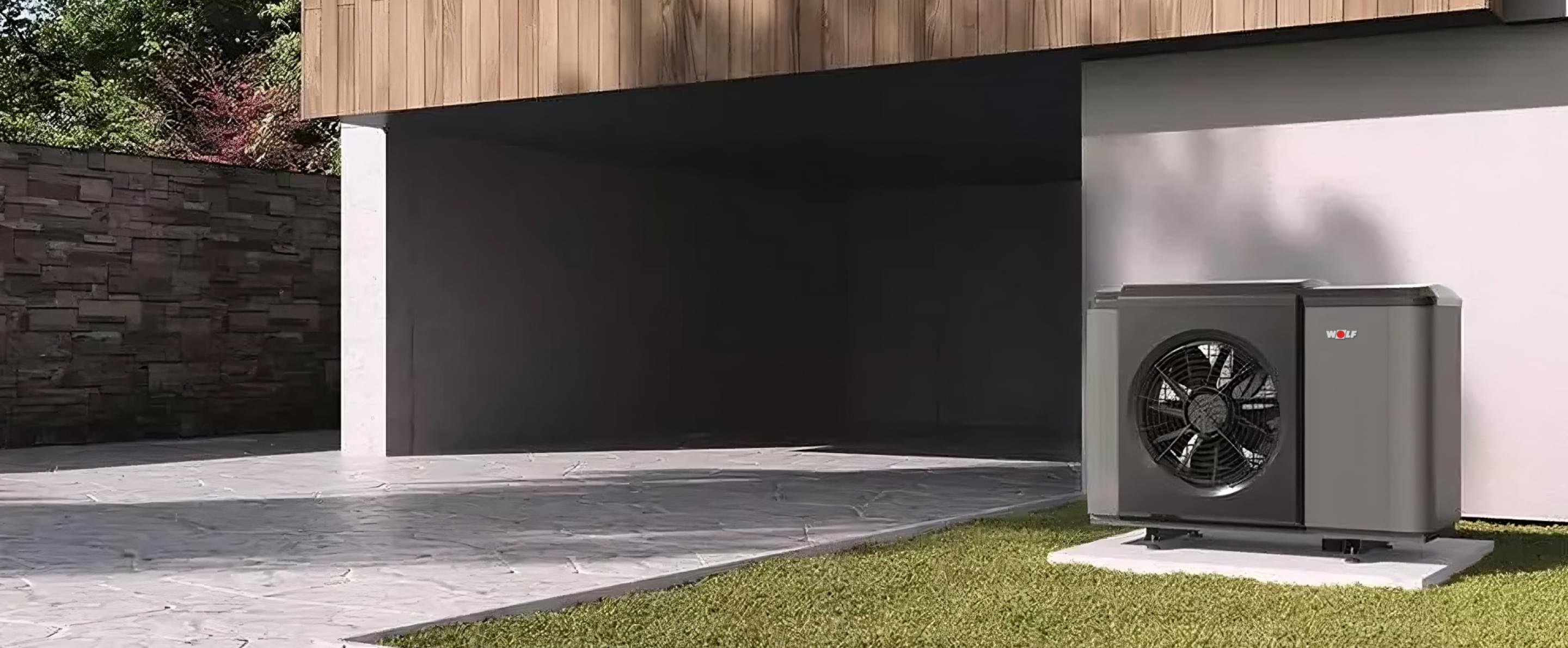 Schwarze WOLF Luft-Wasser-Wärmepumpe auf grünem Rasen vor modernem Gebäude mit Holzverkleidung und Einfahrt.