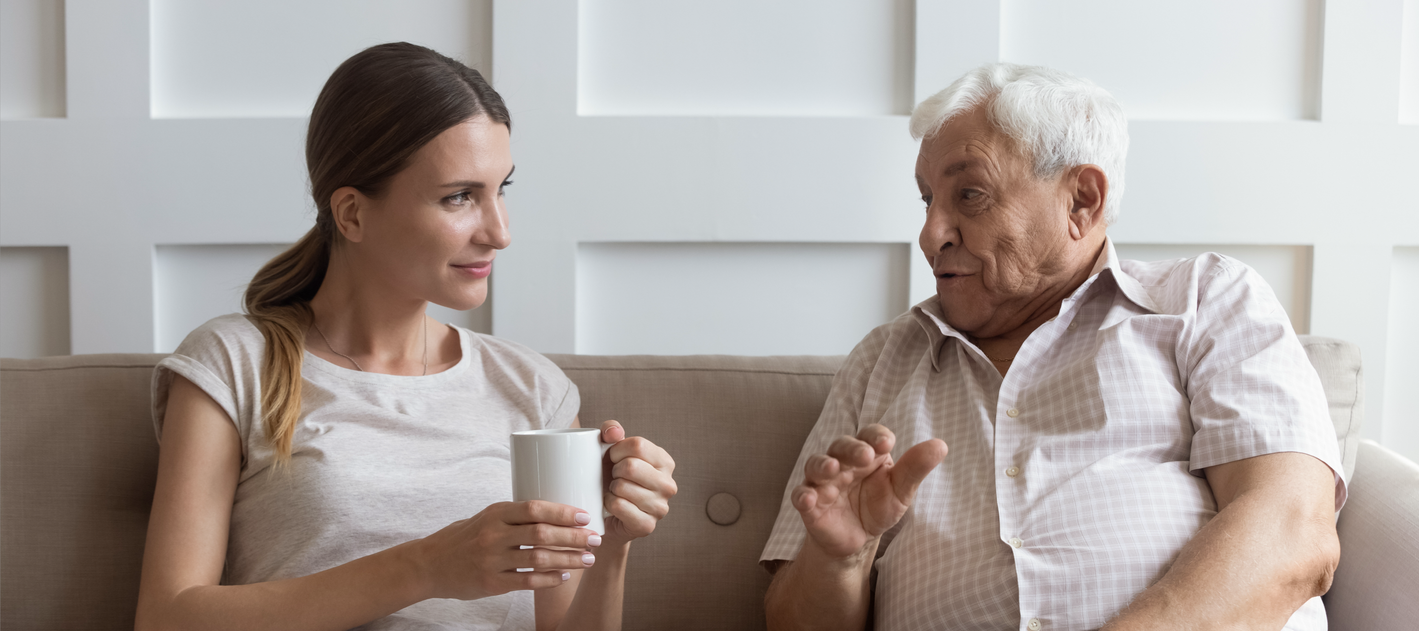 Älterer Mann und junge Frau sitzen auf dem Sofa, unterhalten sich und lächeln, die Frau hält eine Tasse in der Hand.