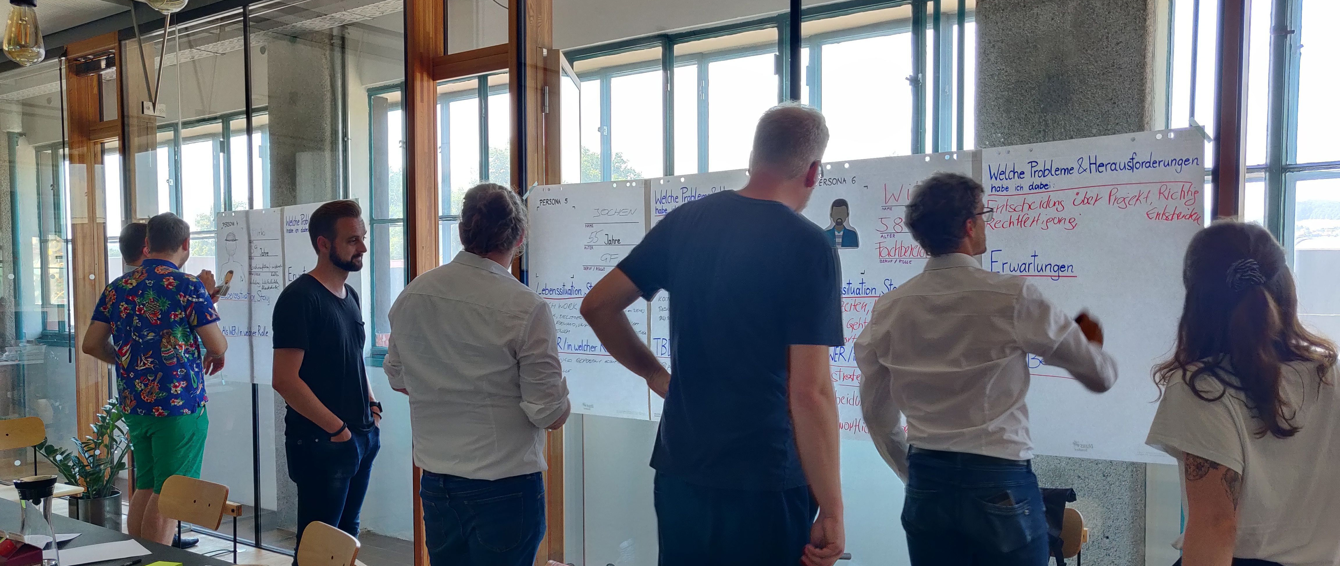 Teilnehmende stehen vor beschrifteten Whiteboards und diskutieren Inhalte in einem lichtdurchfluteten Raum.