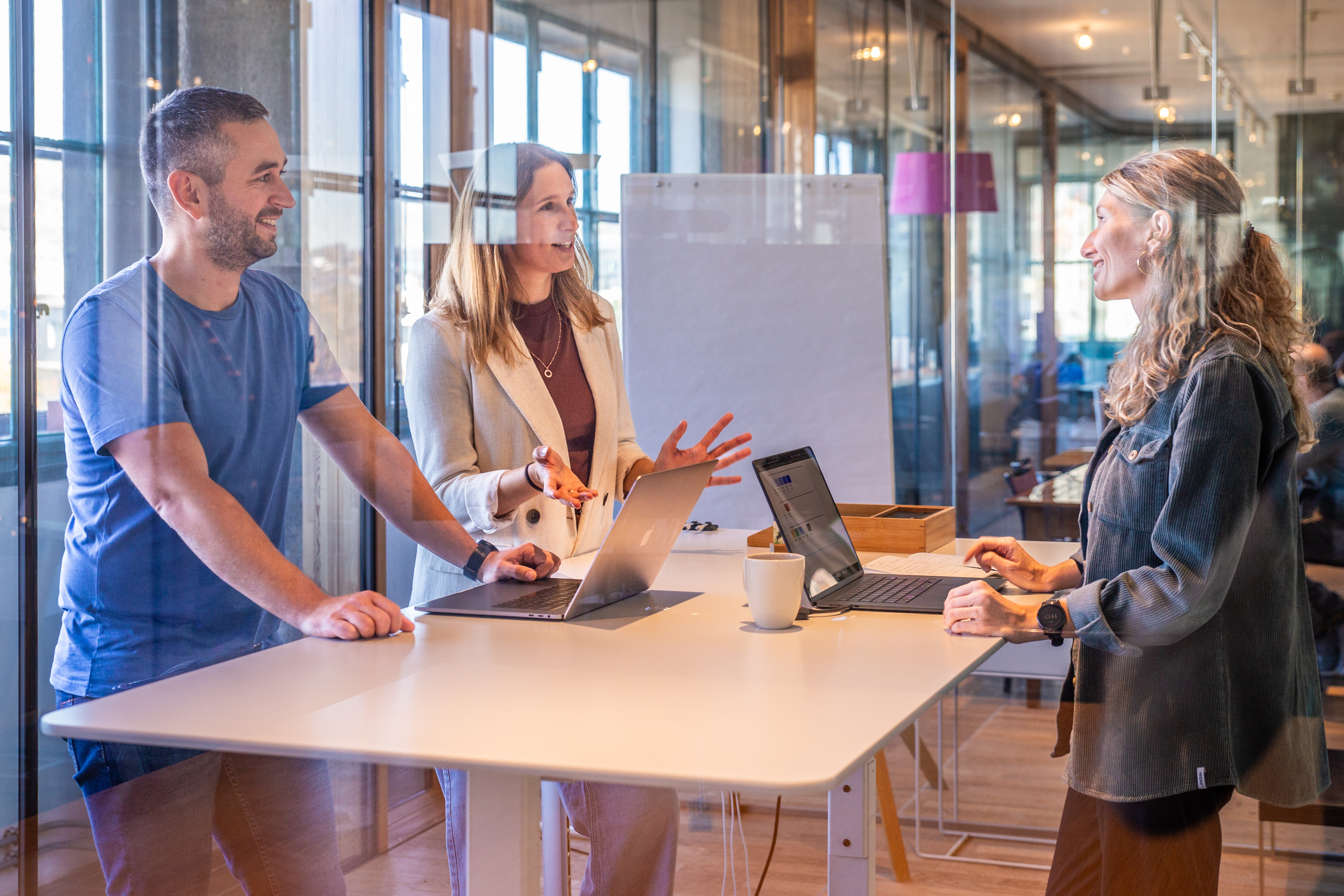 Drei Personen halten eine Besprechung in einem Büro mit Glaswänden ab; Laptops stehen auf dem Tisch, im Hintergrund ist eine Tafel zu sehen.