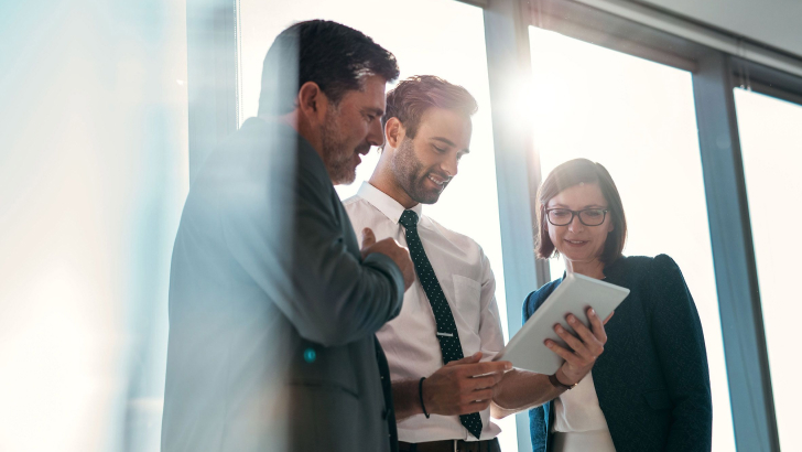 Drei Personen im Business-Outfit stehen im Sonnenlicht und schauen gemeinsam auf ein Tablet.