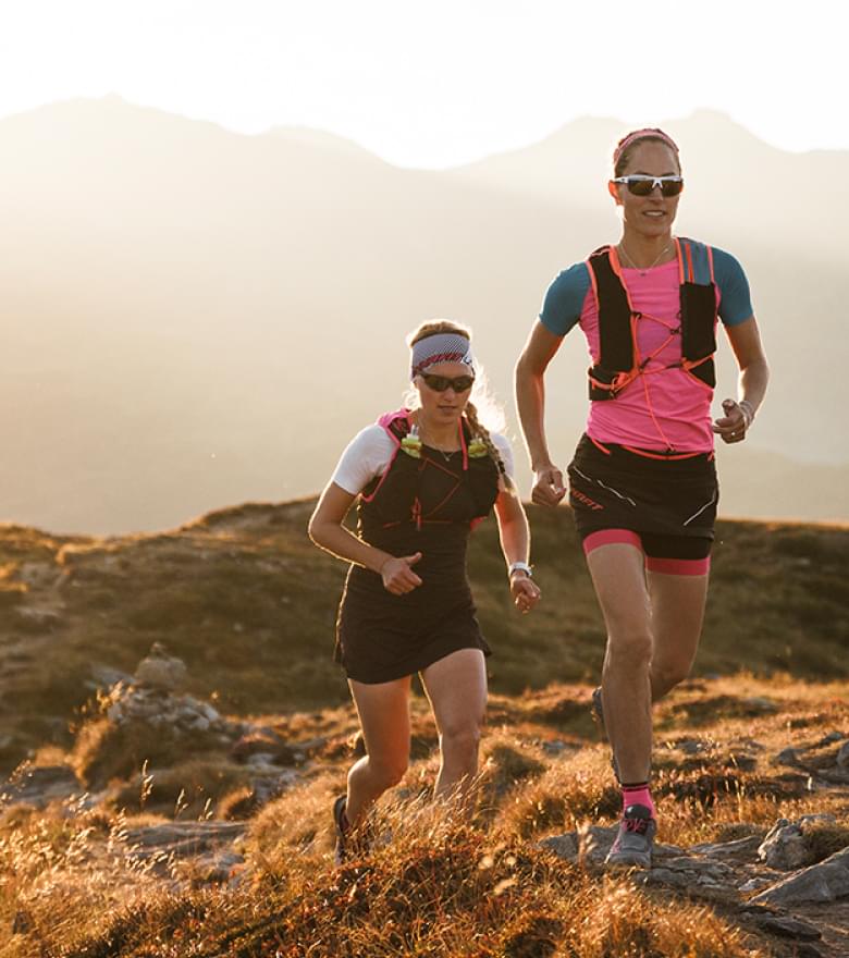 Zwei Frauen mit evil eye Sportbrillen laufen in einer Berglandschaft.