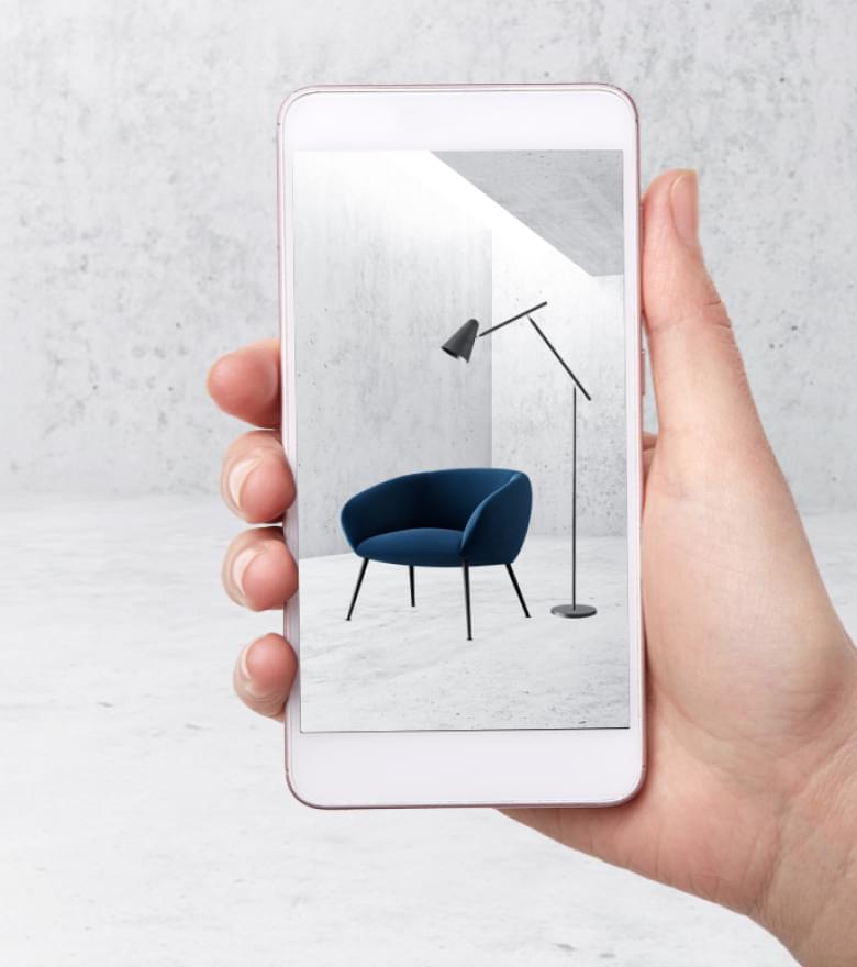 Hand hält Smartphone mit AR-Ansicht eines blauen Sessels und einer Stehlampe in minimalistischem Raum.