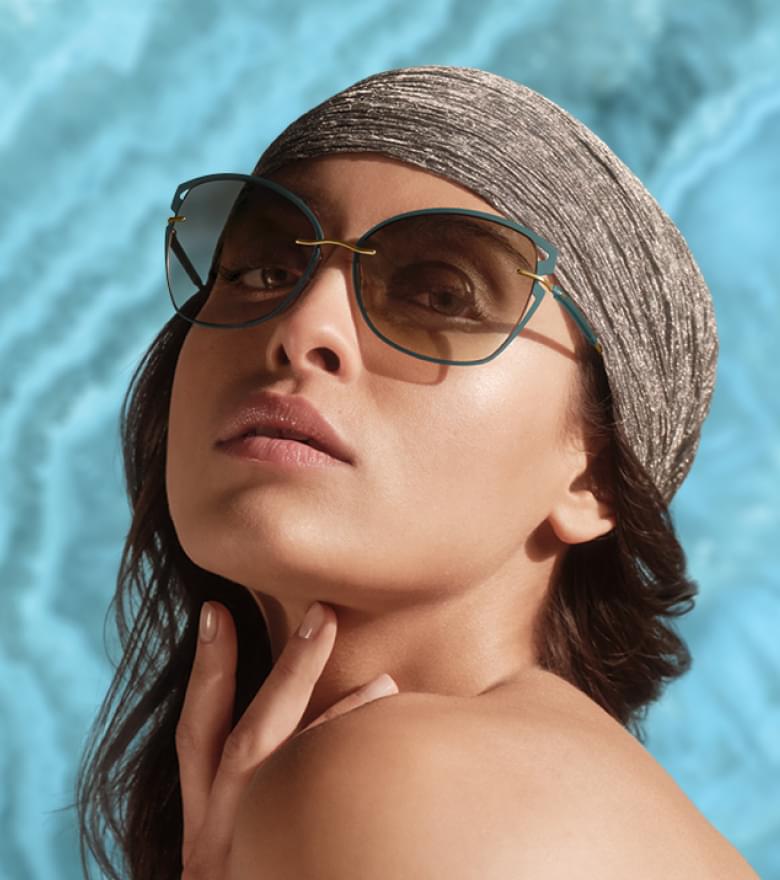 Eine Frau mit Kopftuch und stilvoller Silhouette Sonnenbrille blickt direkt in die Kamera, vor blauem Hintergrund.