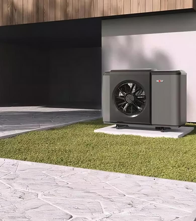 Schwarze WOLF Luft-Wasser-Wärmepumpe auf grünem Rasen vor modernem Gebäude mit Holzverkleidung und Einfahrt.