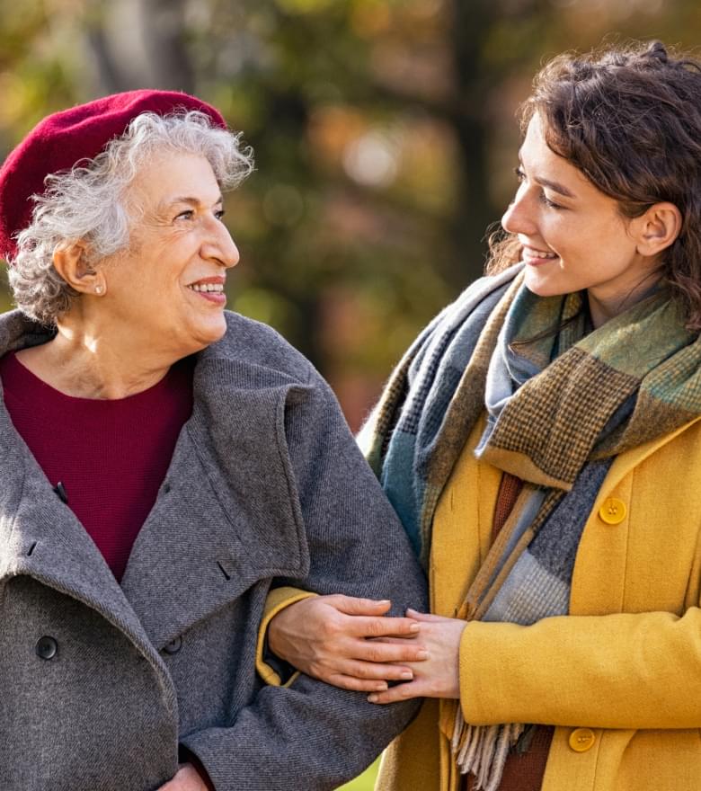 Junge Frau spaziert lachend mit älterer Frau durch herbstlichen Park, beide warm gekleidet und arm in arm.
