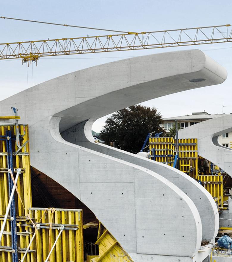 Brückenbau mit geschwungenen Betonelementen und gelbem Kran bei Sonnenschein, im Hintergrund Wohn- und Bürogebäude.