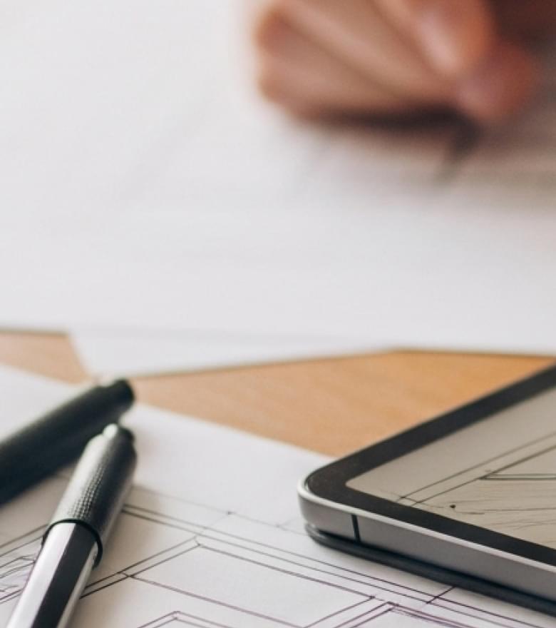 Eine Hand skizziert auf Bauplänen neben einem Tablet mit Raumplanungs-App und Stiften.