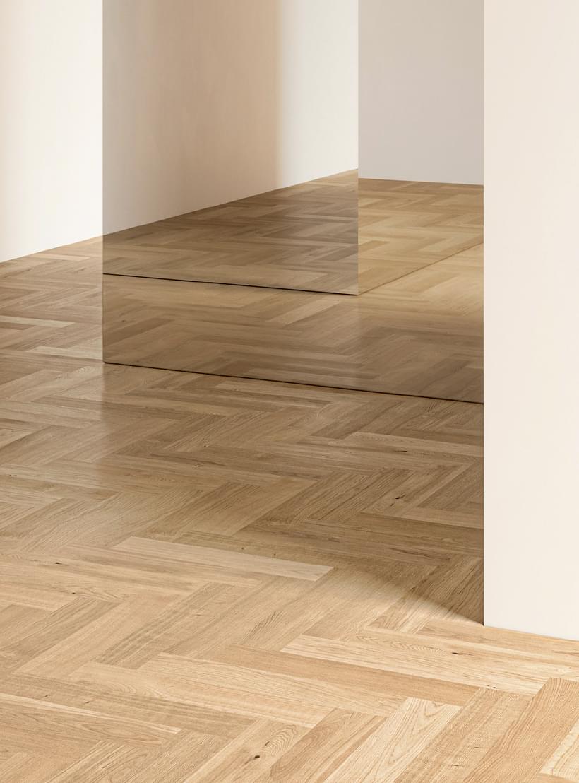 In einem minimalistisch gestalteten Innenraum reflektiert ein heller Fischgrätparkettboden die Spiegelung an einer Wand.