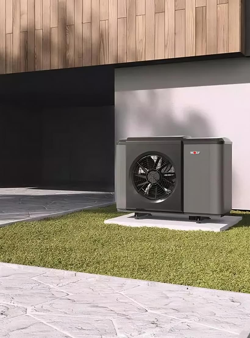 Schwarze WOLF Luft-Wasser-Wärmepumpe auf grünem Rasen vor modernem Gebäude mit Holzverkleidung und Einfahrt.