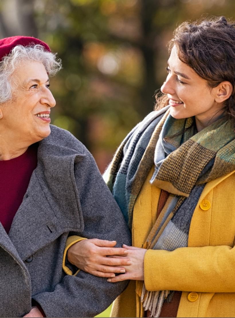 Junge Frau spaziert lachend mit älterer Frau durch herbstlichen Park, beide warm gekleidet und arm in arm.