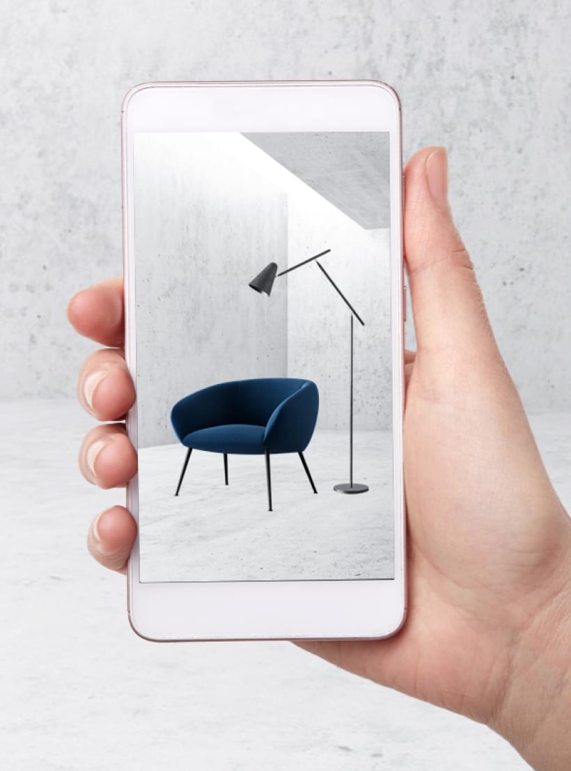 Hand hält Smartphone mit AR-Ansicht eines blauen Sessels und einer Stehlampe in minimalistischem Raum.