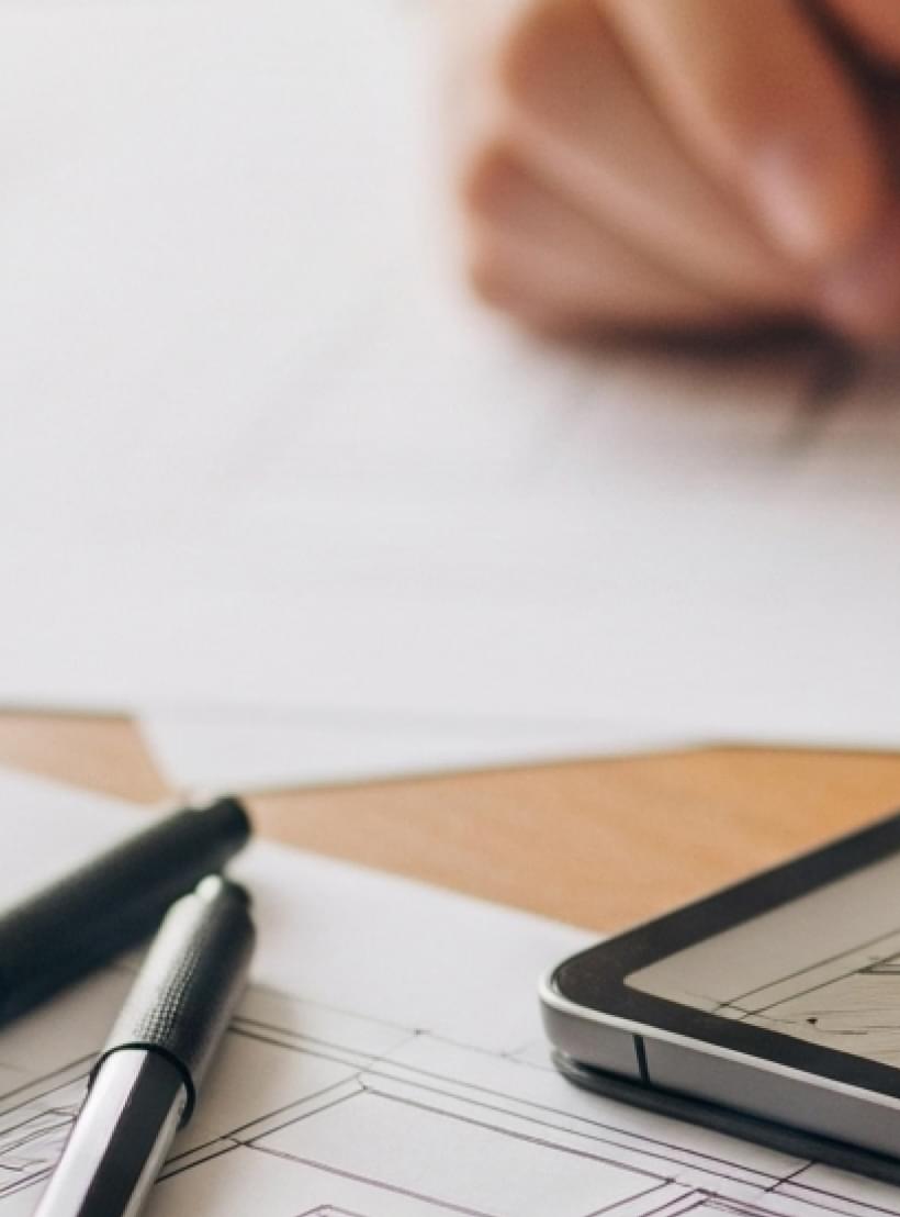 Eine Hand skizziert auf Bauplänen neben einem Tablet mit Raumplanungs-App und Stiften.