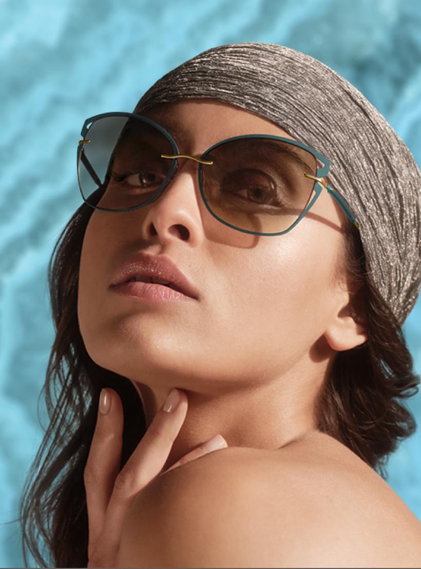 Eine Frau mit Kopftuch und stilvoller Silhouette Sonnenbrille blickt direkt in die Kamera, vor blauem Hintergrund.