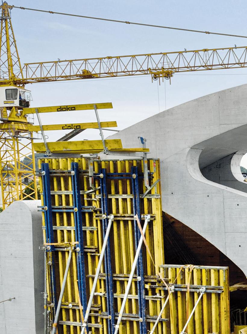 Brückenbau mit geschwungenen Betonelementen und gelbem Kran bei Sonnenschein, im Hintergrund Wohn- und Bürogebäude.
