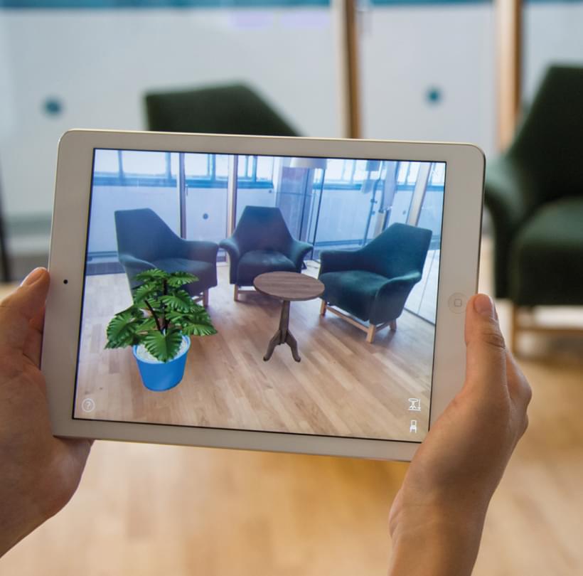 Person hält ein Tablet mit Augmented Reality-Ansicht von Möbeln und Pflanze in einem Raum.